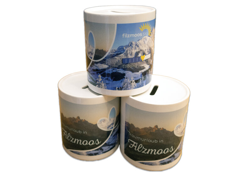 Drei zylindrische Filzmoos-Spardosen mit Bergpanorama und Schlitz im Deckel vor hellem Hintergrund. | © Bergbahnen Filzmoos GmbH