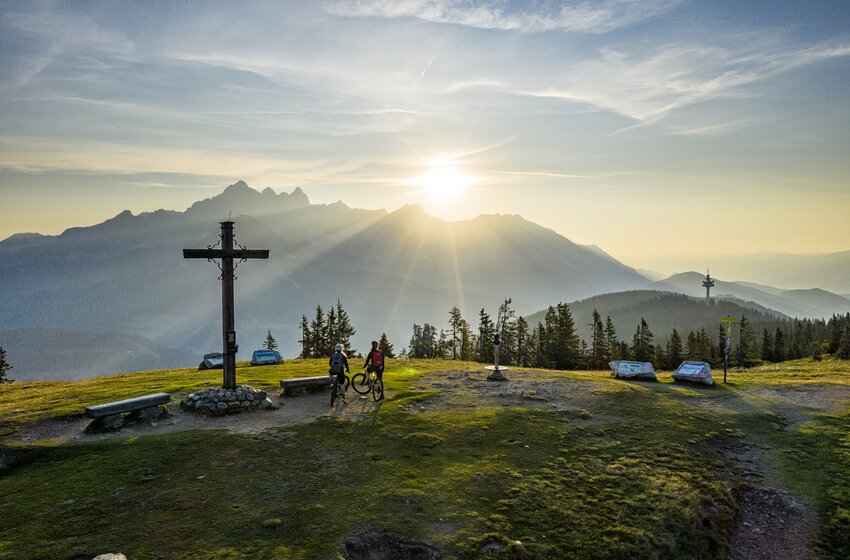 Two mountain bikers stand with bikes at a summit cross on grass, sun over mountain range, forest and distant view | © Bergbahnen Filzmoos GmbH