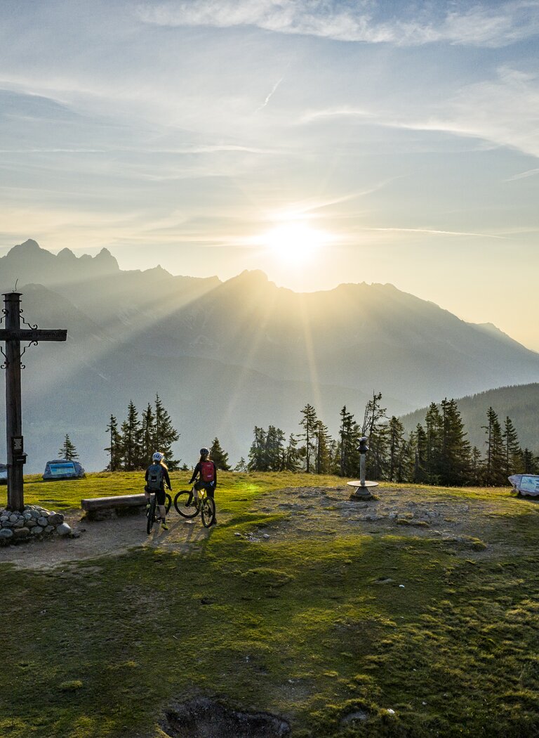 Two mountain bikers stand with bikes at a summit cross on grass, sun over mountain range, forest and distant view | © Bergbahnen Filzmoos GmbH