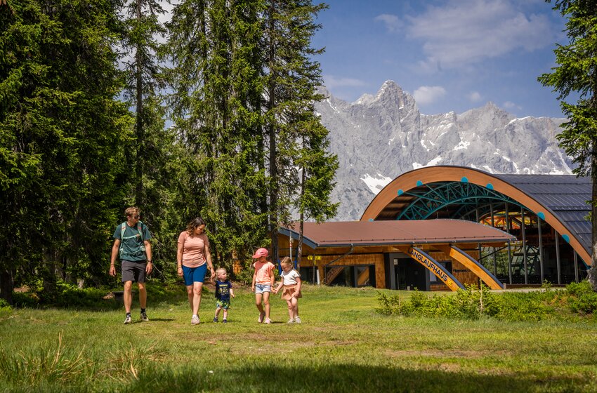 Two adults and three children walk across grass with mountain backdrop beside modern wooden building near forest | © Bergbahnen Filzmoos GmbH