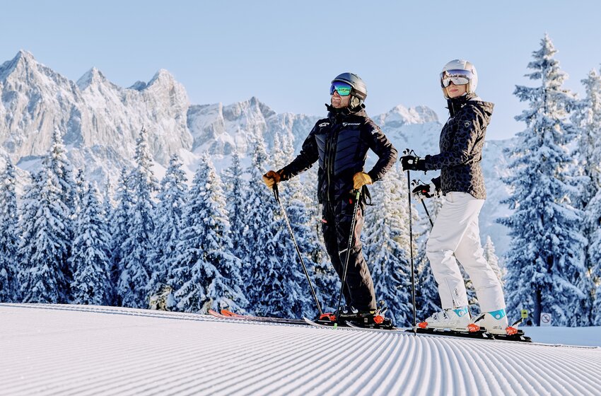 Two skiers stand on groomed slope with snow-covered trees and Dachstein mountains in background. | © Bergbahnen Filzmoos GmbH