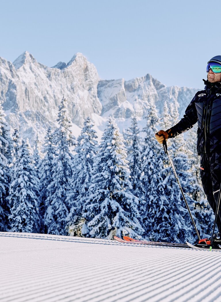 Two skiers stand on groomed slope with snow-covered trees and Dachstein mountains in background. | © Bergbahnen Filzmoos GmbH