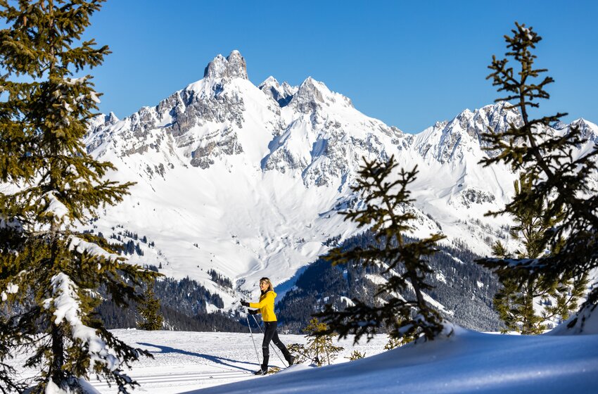 Woman in yellow outfit skis cross-country through snow with mountain scenery and fir trees visible | © Bergbahnen Filzmoos GmbH