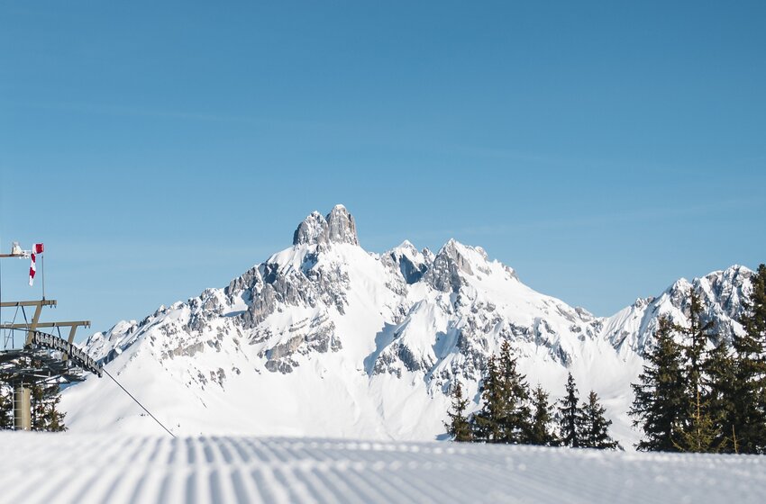 Frisch präparierte Skipiste mit Blick auf schneebedeckte Gipfel | © Bergbahnen Filzmoos GmbH Blick auf präparierte Piste, verschneite Bergspitzen, Skiliftstation und Tannen im Vordergrund | © Bergbahnen Filzmoos GmbH