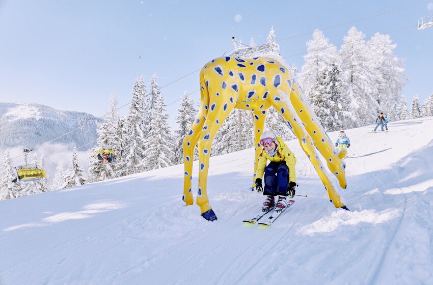 A smiling child skis through a yellow giraffe figure with snowy landscape and trees behind. | © Bergbahnen Filzmoos GmbH