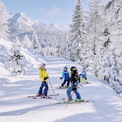 Four kids stand on a snowy forest slope, surrounded by trees and mountain scenery in winter. | © Bergbahnen Filzmoos GmbH