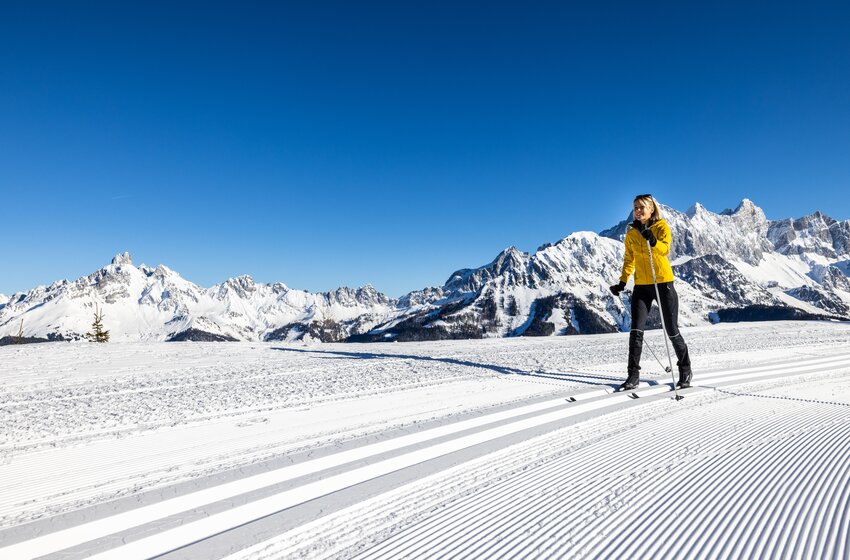 Woman in yellow jacket skis on freshly groomed track with snowy alpine mountains behind her. | © Bergbahnen Filzmoos GmbH