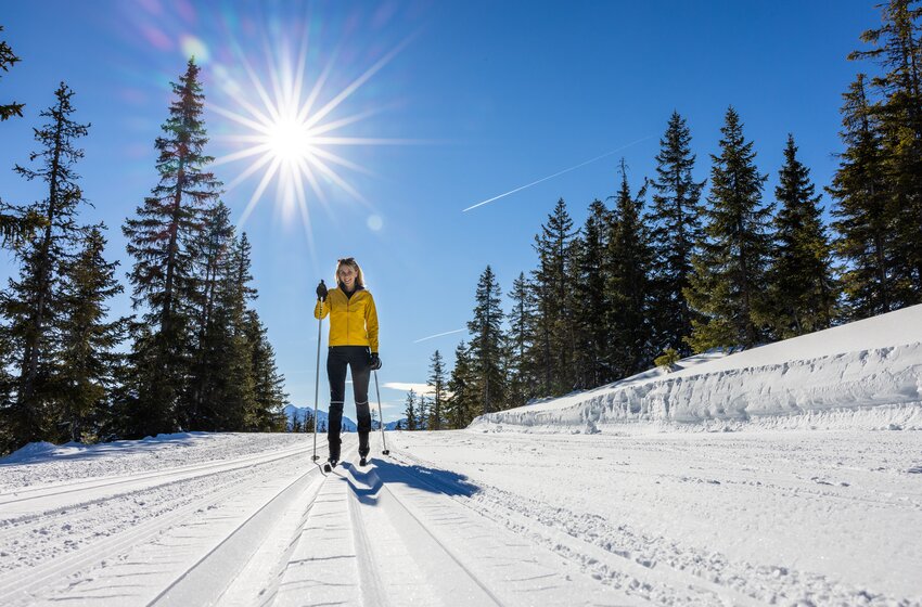 Woman in yellow jacket skis on groomed trail through snowy forest in bright winter sunshine. | © Bergbahnen Filzmoos GmbH