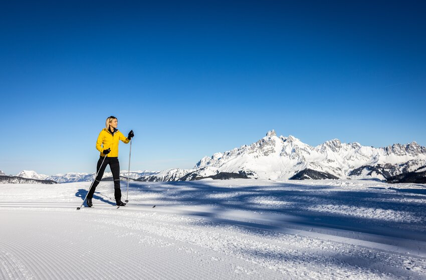 Woman in yellow jacket cross-country skiing on groomed trail with snow-covered mountain view. | © Bergbahnen Filzmoos GmbH