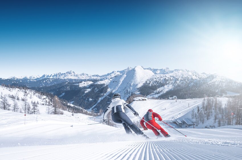 Two skiers carving down a sunlit, freshly groomed slope with panoramic views of the snowy mountains in Ski amadé. | © Ski amadé GmbH